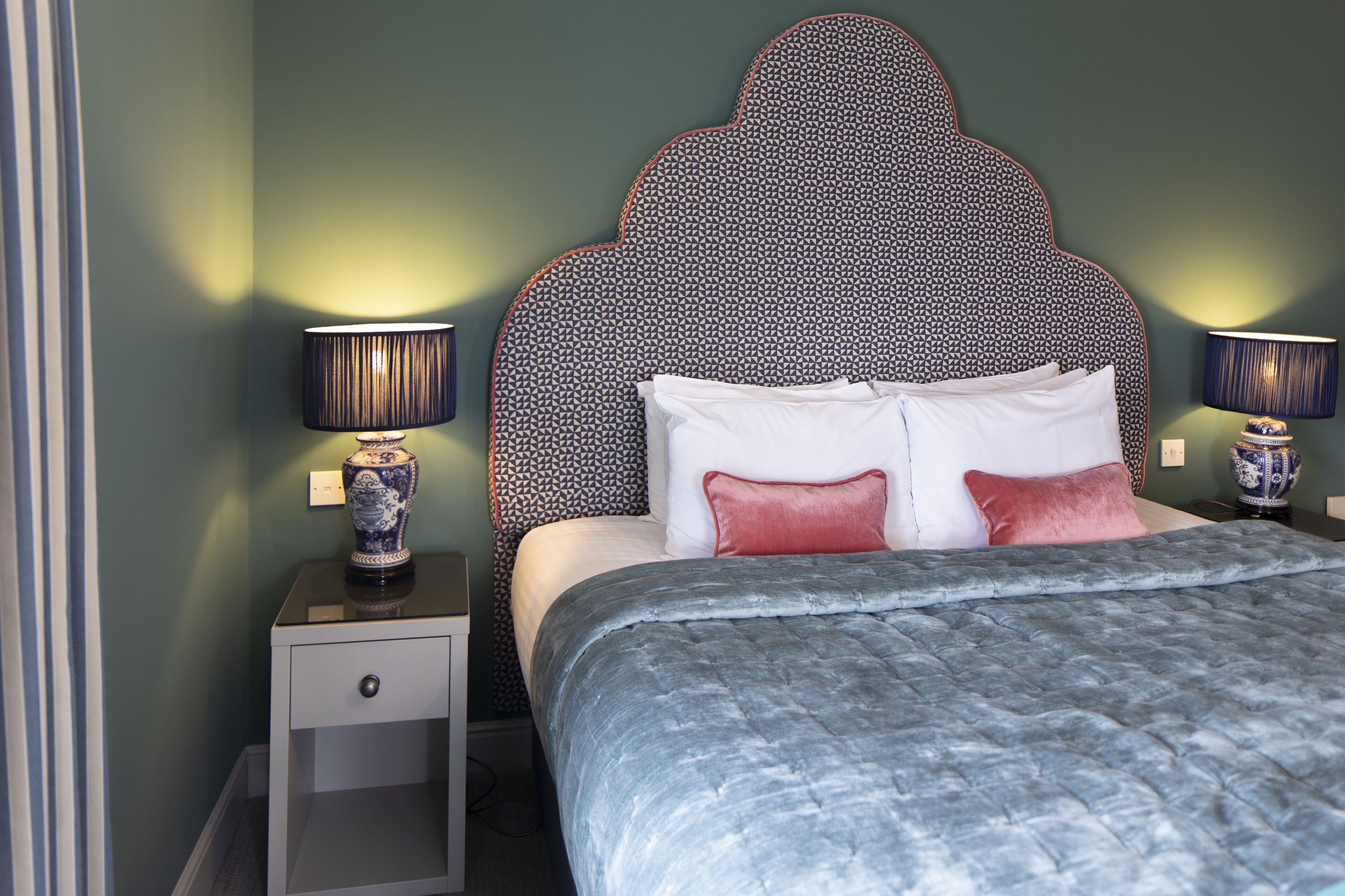 A bedroom with a patterned headboard, two bedside tables with lamps, a bed with a grey quilt, and a small bedside drawer.