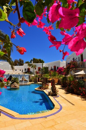 Een zwembad omringd door witte gebouwen, palmbomen en felroze bougainvillea's.