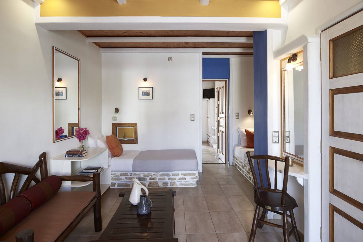 Un salon bien éclairé, avec un canapé, une table basse et un fauteuil. On y trouve une cheminée avec un manteau en pierre, un plafond en bois et un couloir qui mène à une autre pièce.