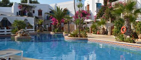 Un área de piscina con edificios blancos, palmeras y flores rosadas.