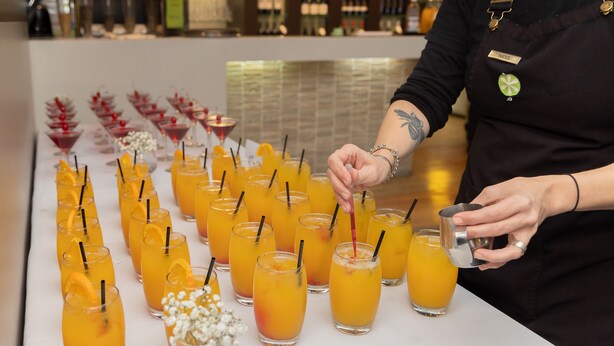 A bar area with multiple glasses of orange juice and cocktails, a bartender preparing drinks, and a bar counter with various bottles and glasses in the background. - Dog Rock Motel (Mount Clarence)