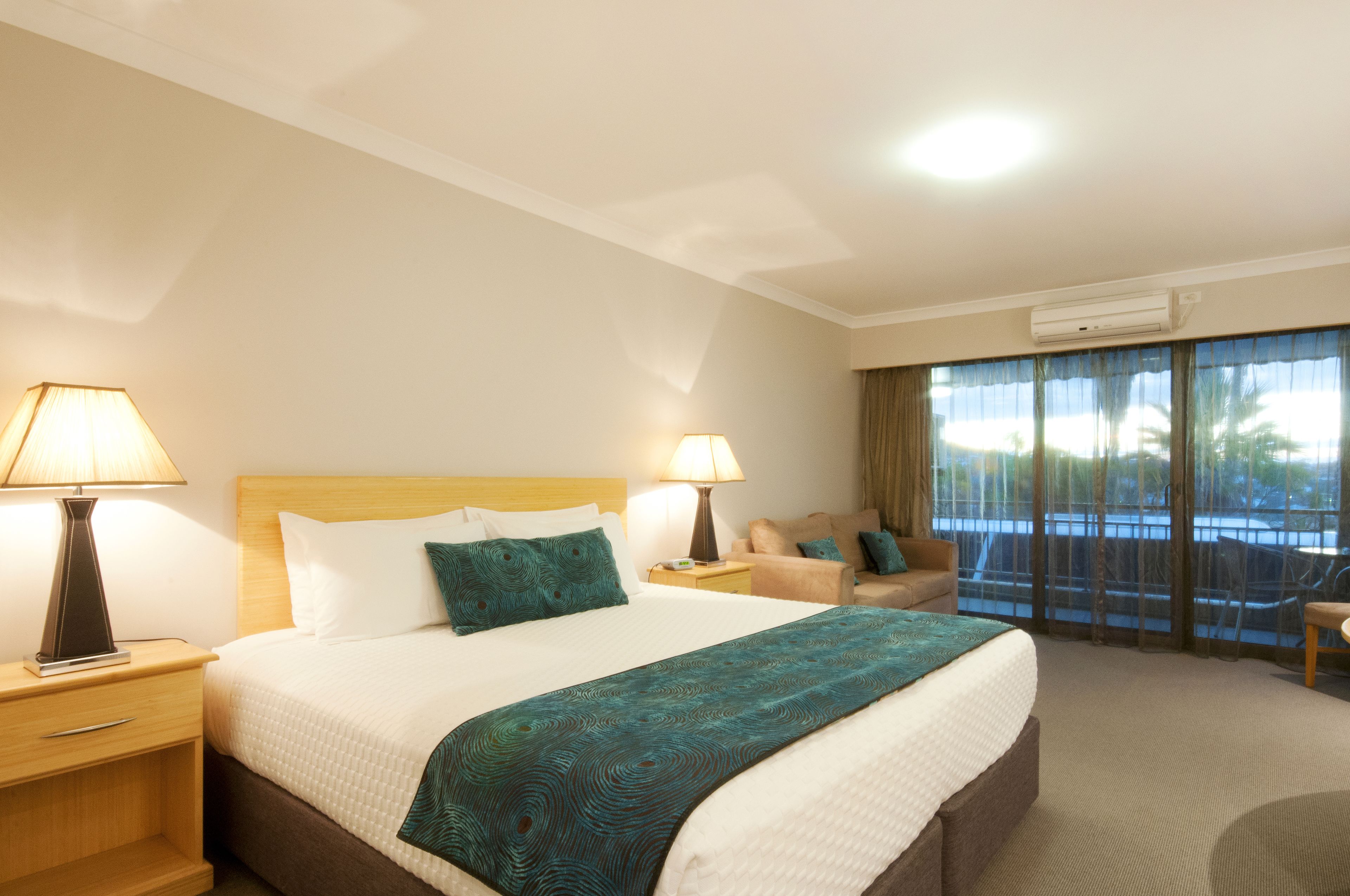 Habitación de hotel con una cama grande, dos mesitas de noche con lámparas, un sofá y un balcón con vistas.