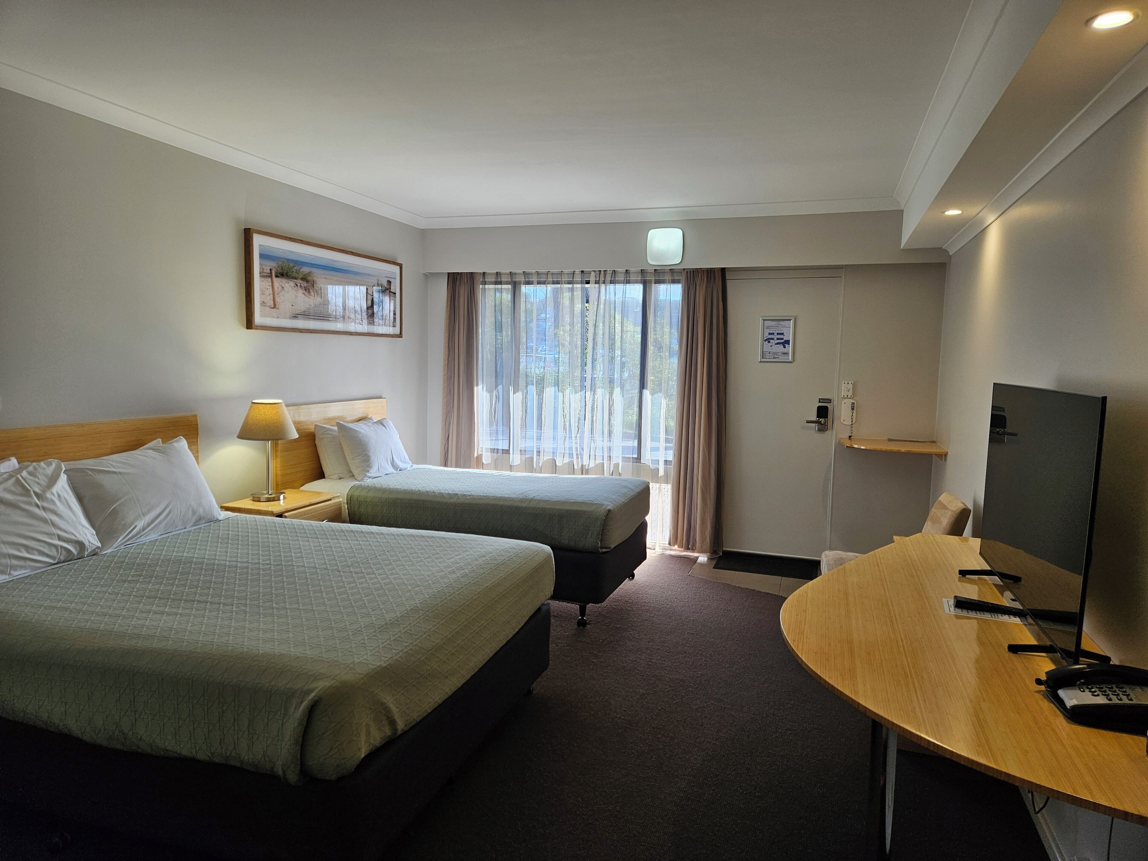 Et hotelværelse med to senge, et skrivebord, et fjernsyn og et vindue med gardiner.