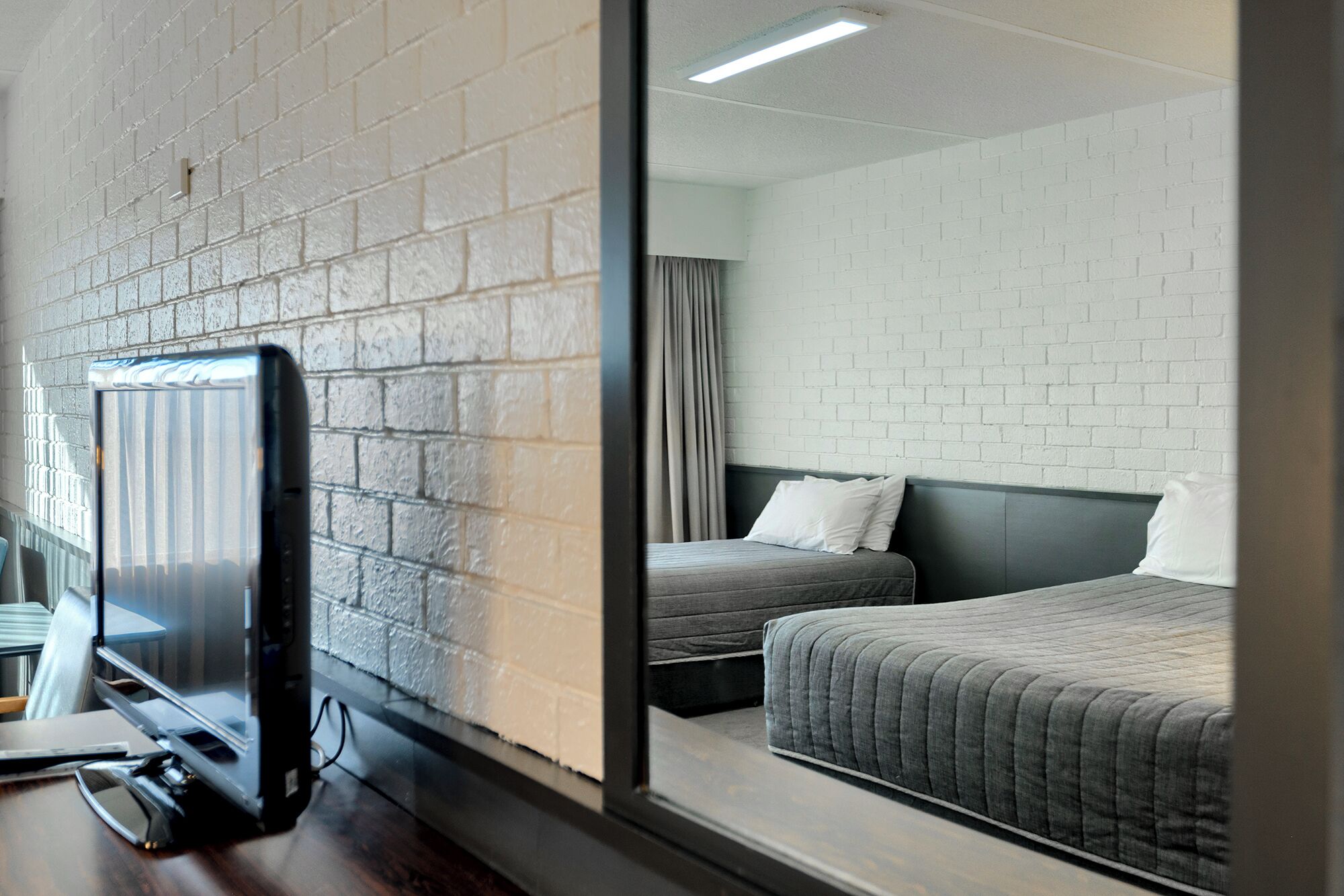 A hotel room with a brick wall, a bed with a grey quilt, a second bed, a TV on a wooden desk, and a window with curtains.