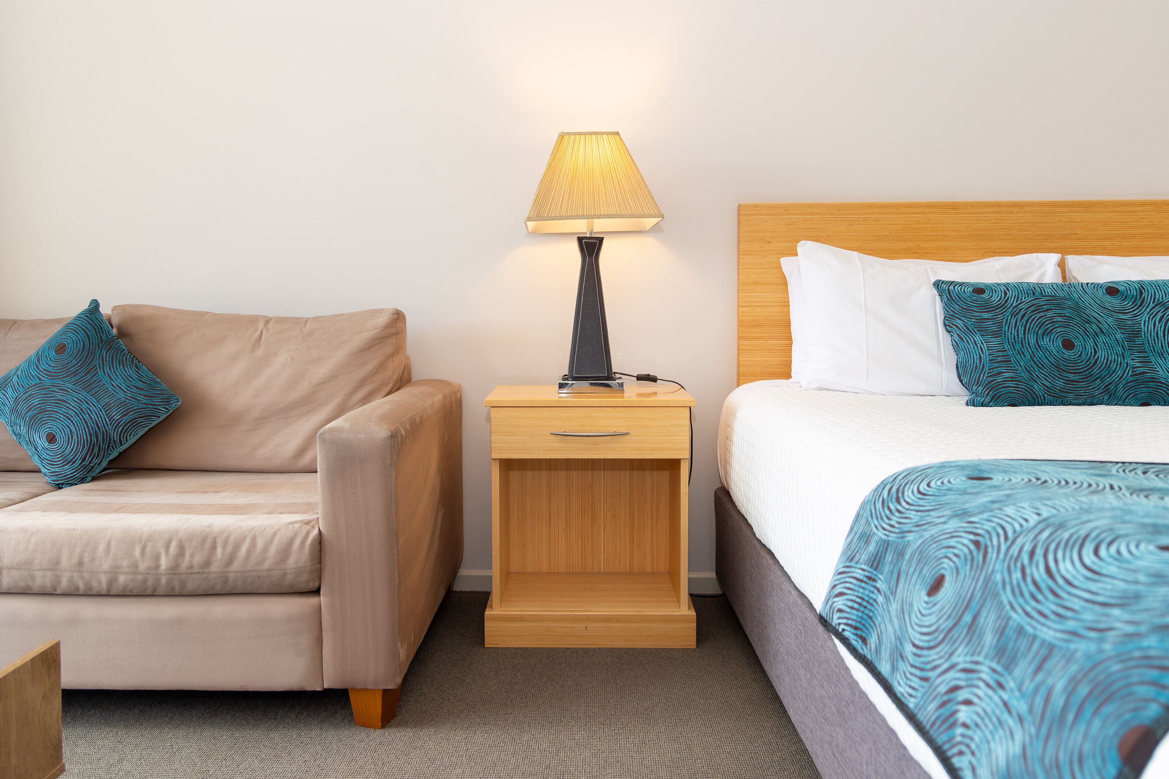A hotel room with a bed, a nightstand with a lamp, a sofa, and a patterned throw blanket.