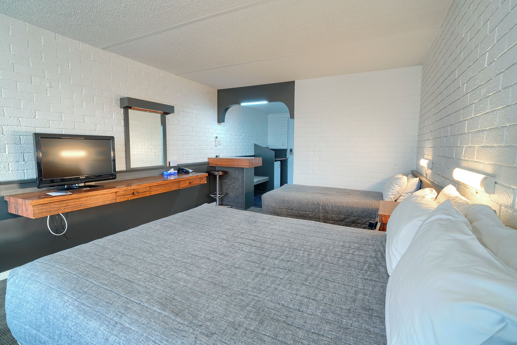 A modern hotel room with a flat-screen TV, a wooden desk, a bar stool, a bed, and a large mirror.