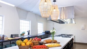 A modern kitchen with a long island, various fruits, pastries, and a decorative light fixture. - Stoke Beach House - Hostel (Manly)