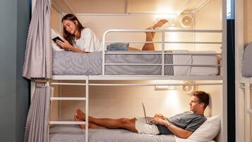A bunk bed room with a person on the top bunk using a smartphone, another person on the bottom bunk using a laptop, and a third person lying on the floor.