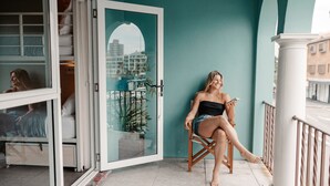 A woman sitting on a wooden chair outside a building with a teal wall, a glass door, and a view of a cityscape. - Stoke Beach House - Hostel (Manly)