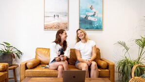 Two people sitting on a brown leather couch with a laptop in front of them, surrounded by potted plants and framed pictures on the wall. - Stoke Beach House - Hostel (Manly)