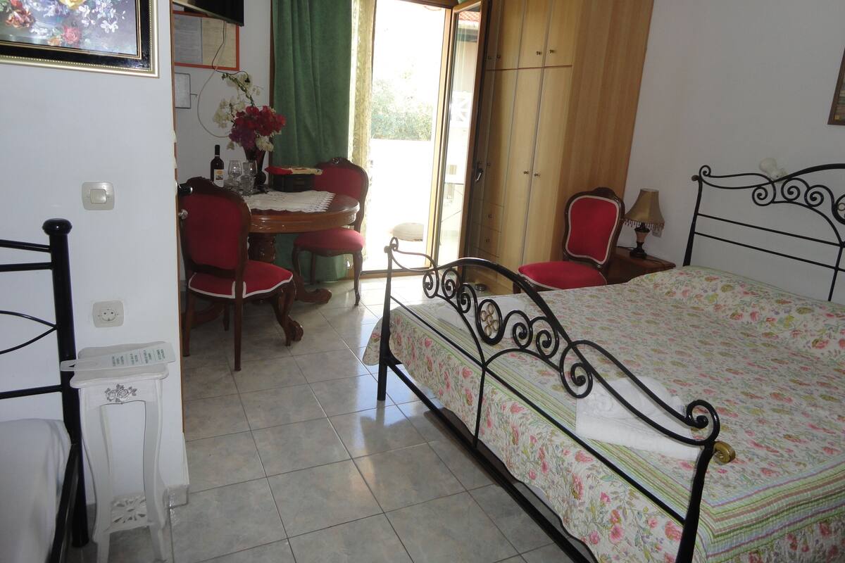 A bedroom with a bed, a desk, two chairs, a table with a vase of flowers, and a wardrobe.