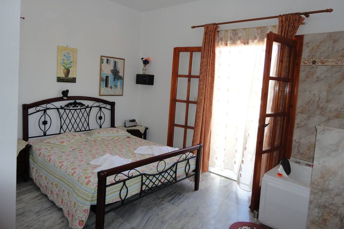 A bedroom with a bed, a nightstand, a window with curtains, and a bathroom with a bathtub.