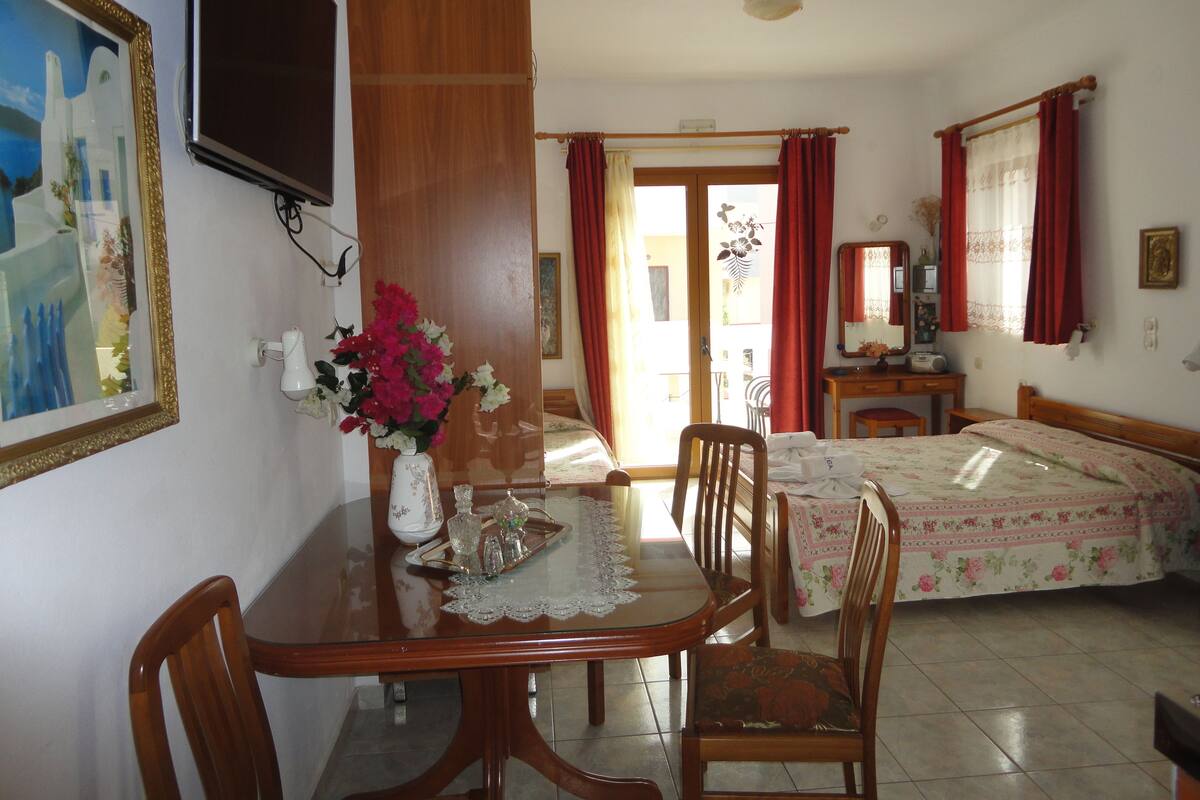 A hotel room with a wooden dining table, chairs, a bed with floral bedding, a TV, and a bathroom with a shower and toilet.