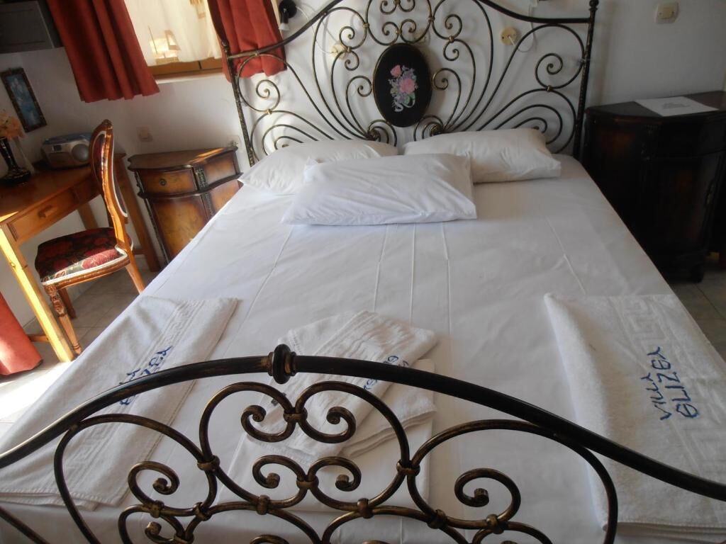 Una cama bien hecha con cabecera metálica, una silla de madera y una mesita con un radio.