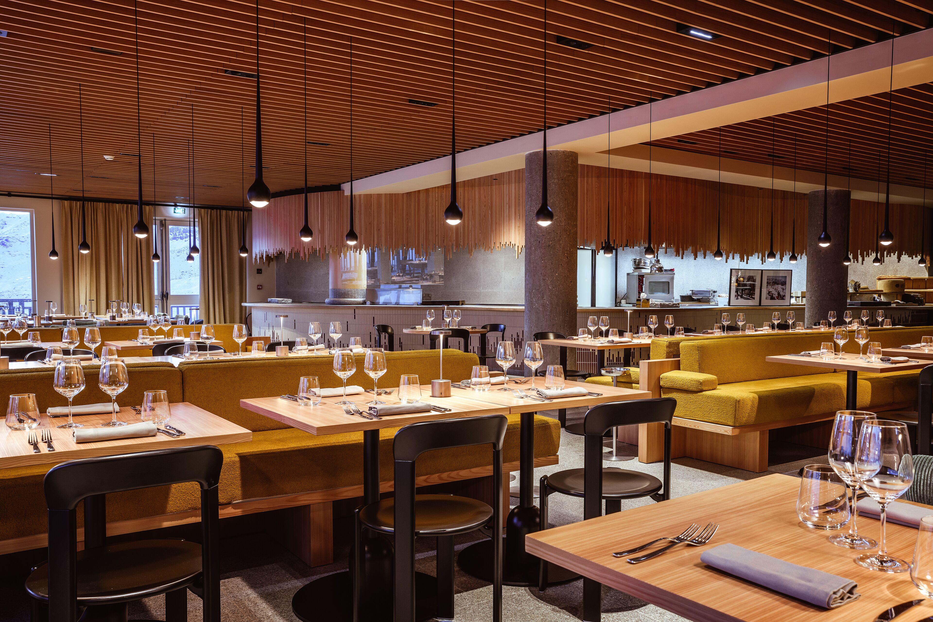 A modern restaurant with wooden accents, multiple dining areas, and a visible kitchen in the background.