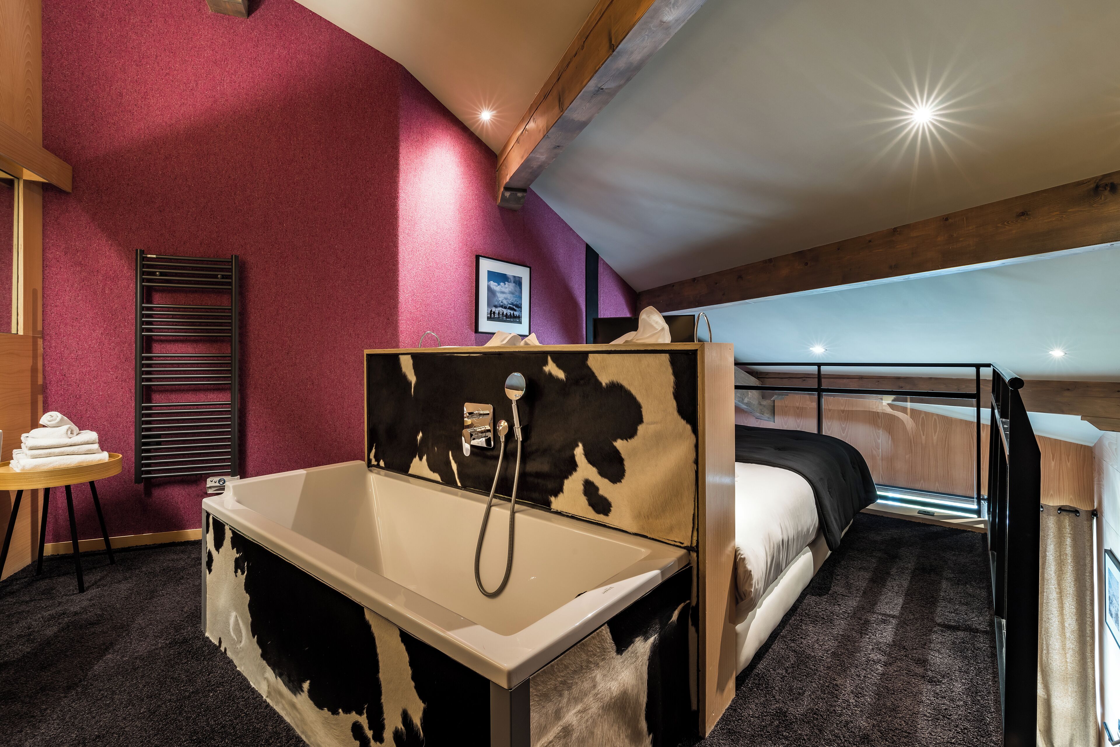 A room with a bed, a small table with towels, a shower, and a cowhide-patterned bench.