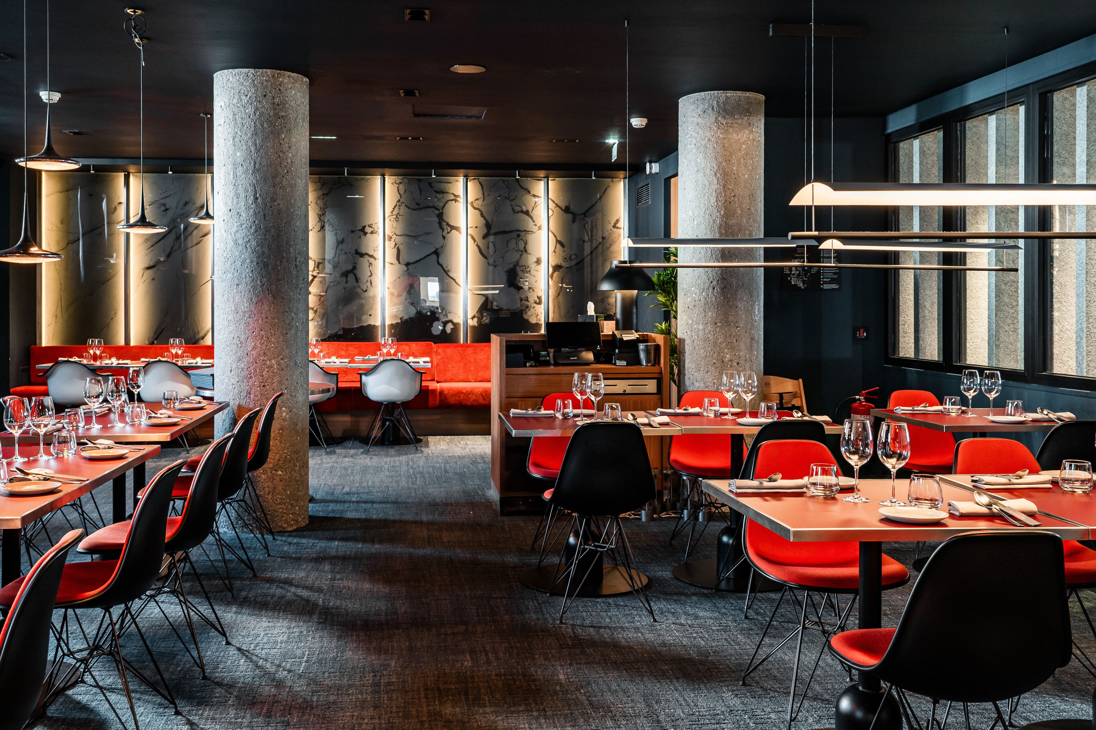 Un intérieur de restaurant moderne, caractérisé par de hauts plafonds, des murs en briques apparentes et un mélange de chaises rouges et noires.