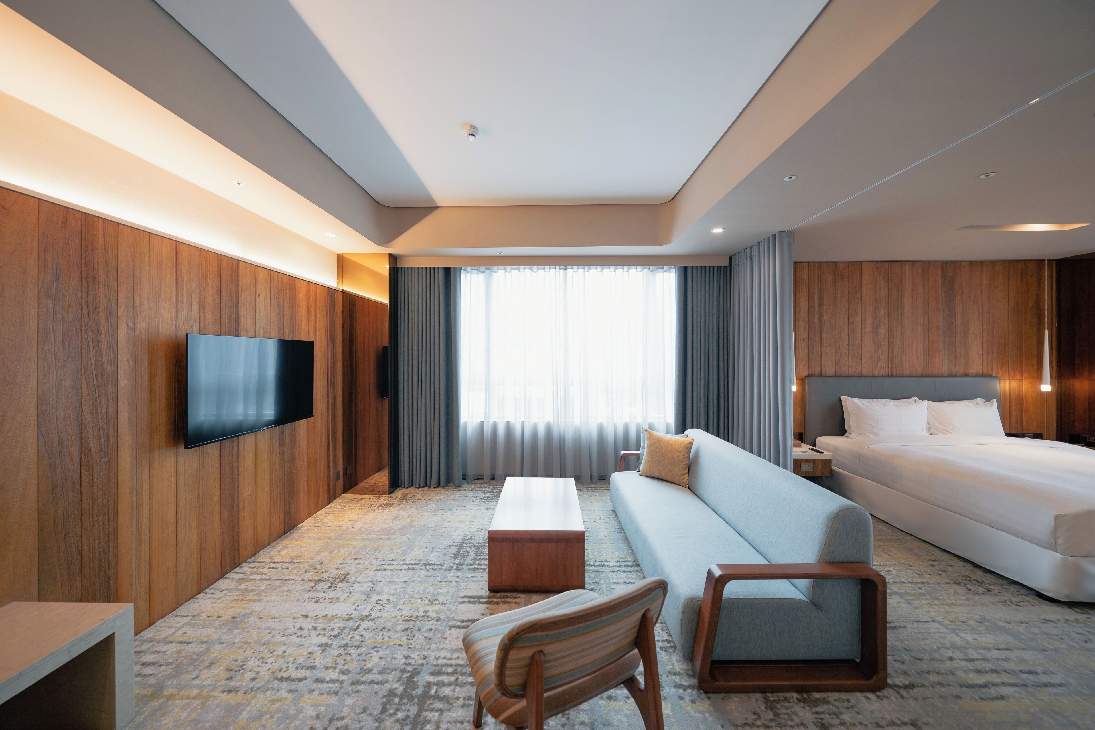 Habitación de hotel con una televisión de pantalla plana montada en la pared, un sofá, una cama, una pequeña mesa de madera, una silla y un ventanal con cortinas.