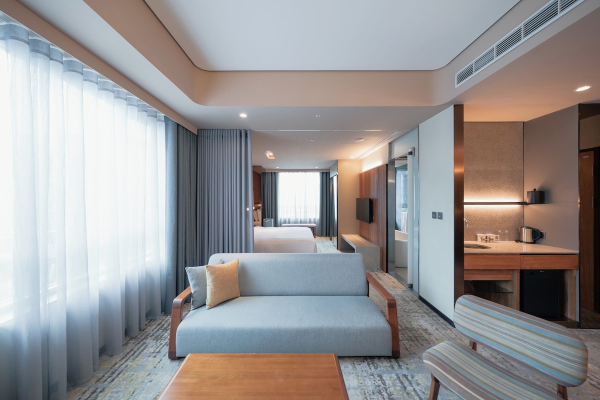 A hotel room with a sofa, a wooden coffee table, a striped chair, a flat-screen TV, a minibar, and a bathroom with a shower and toilet.
