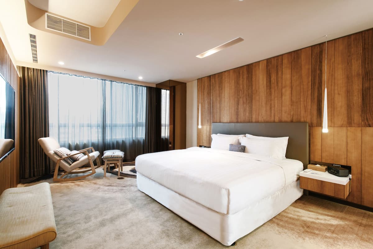 Une chambre d’hôtel moderne avec un grand lit, un coin salon comprenant un fauteuil et un pouf, un mur décoratif en bois et une vue par la fenêtre.