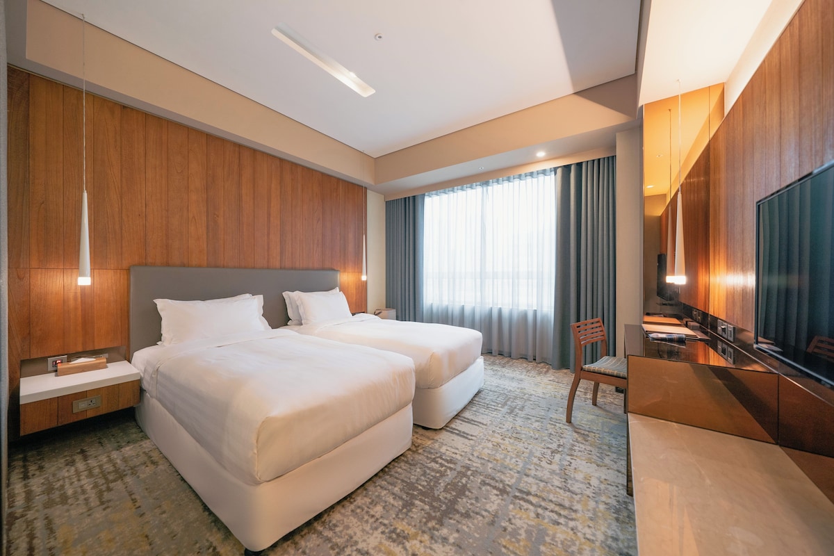 A hotel room with two beds, a desk, a chair, a television, and a large window with curtains.