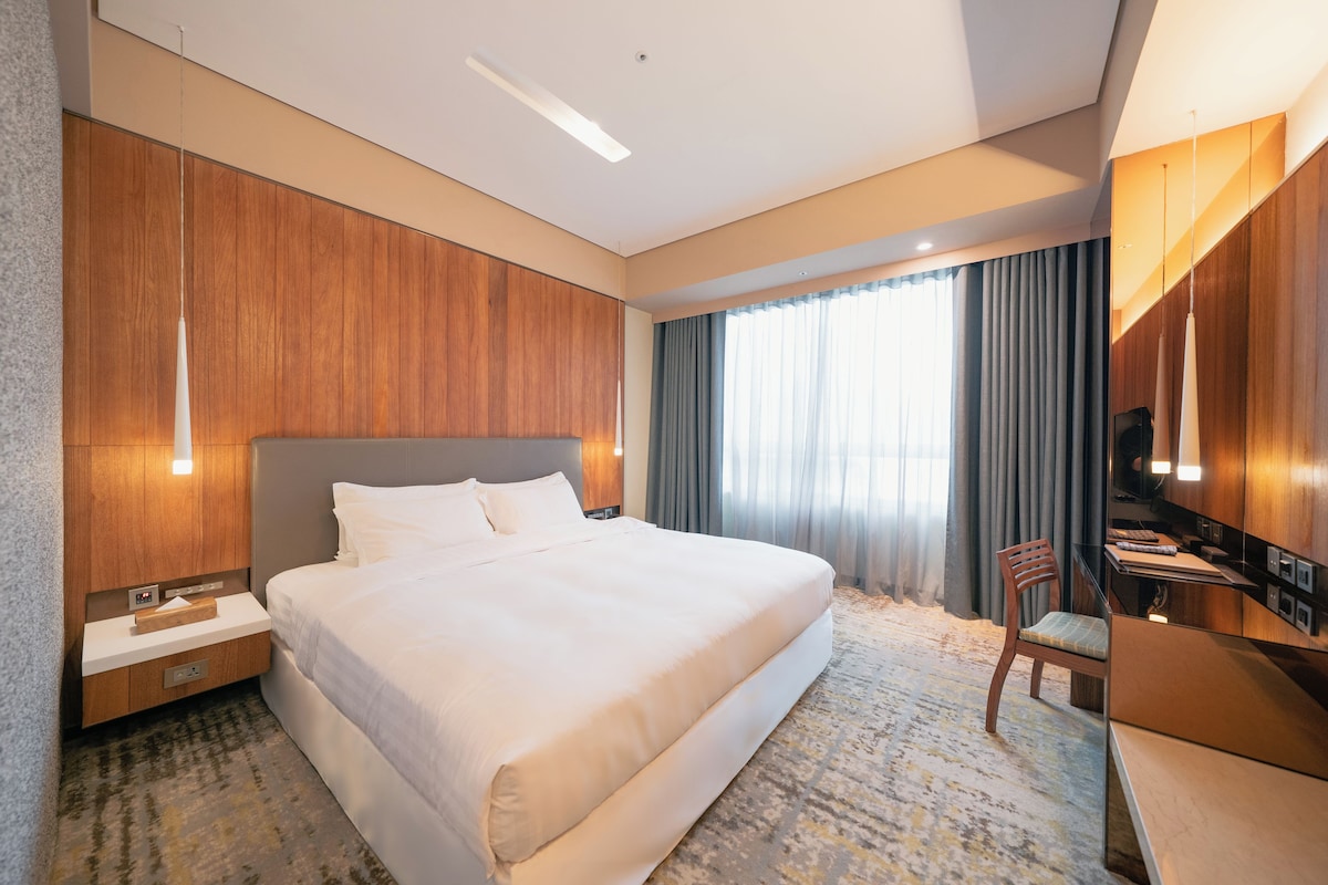 A hotel room with a large bed, a desk with a chair, a television, and a window with curtains.
