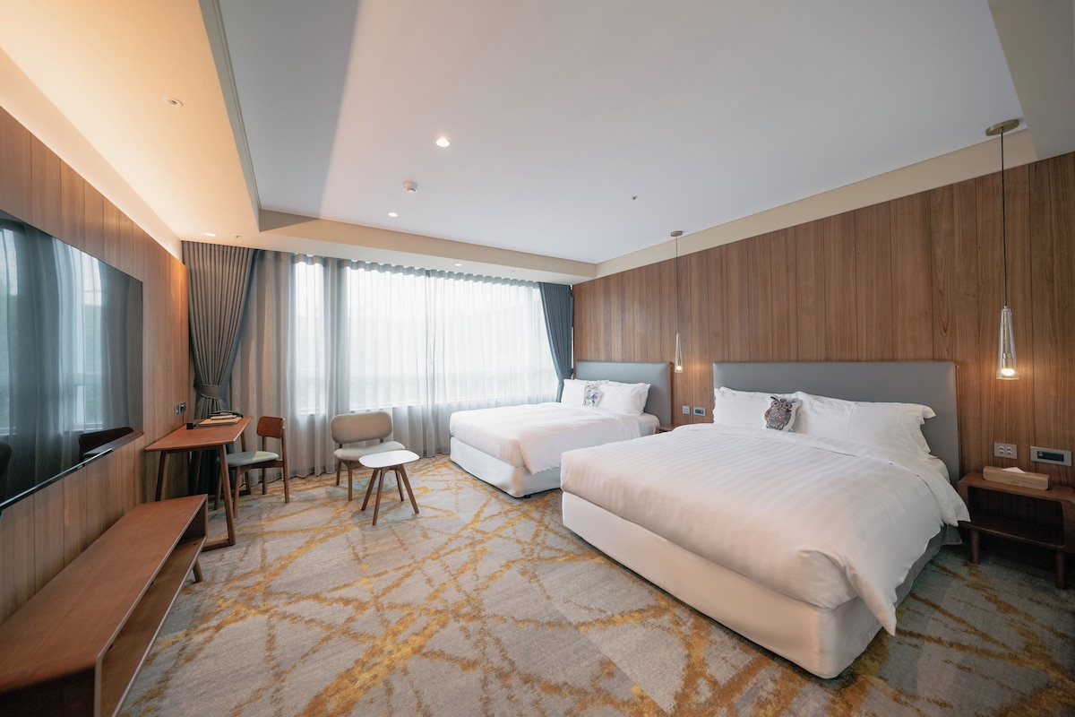 A hotel room with two beds, a desk, a chair, a TV, and a large window with curtains.