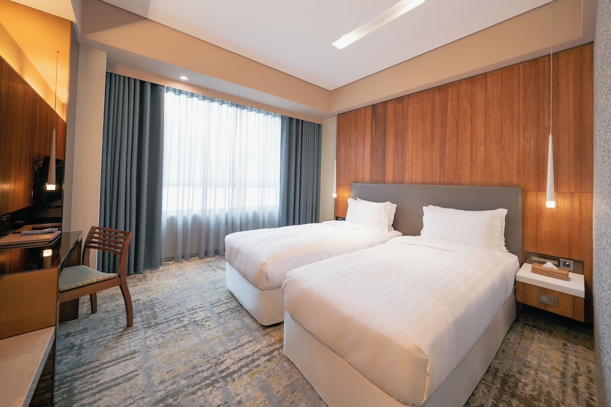 A hotel room with two beds, a desk, a chair, and a large window with curtains.