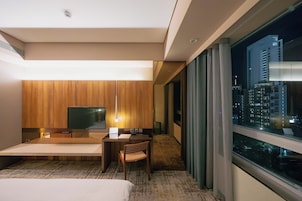 Een hotelkamer met een bed, een bureau met stoel, een televisie en uitzicht op een nachtzicht van de stad.