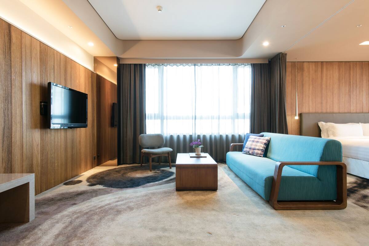 Une chambre d’hôtel moderne avec un mur d’accent en bois, un téléviseur à écran plat fixé au-dessus d’une table basse en bois, un canapé bleu et une grande fenêtre avec des rideaux.