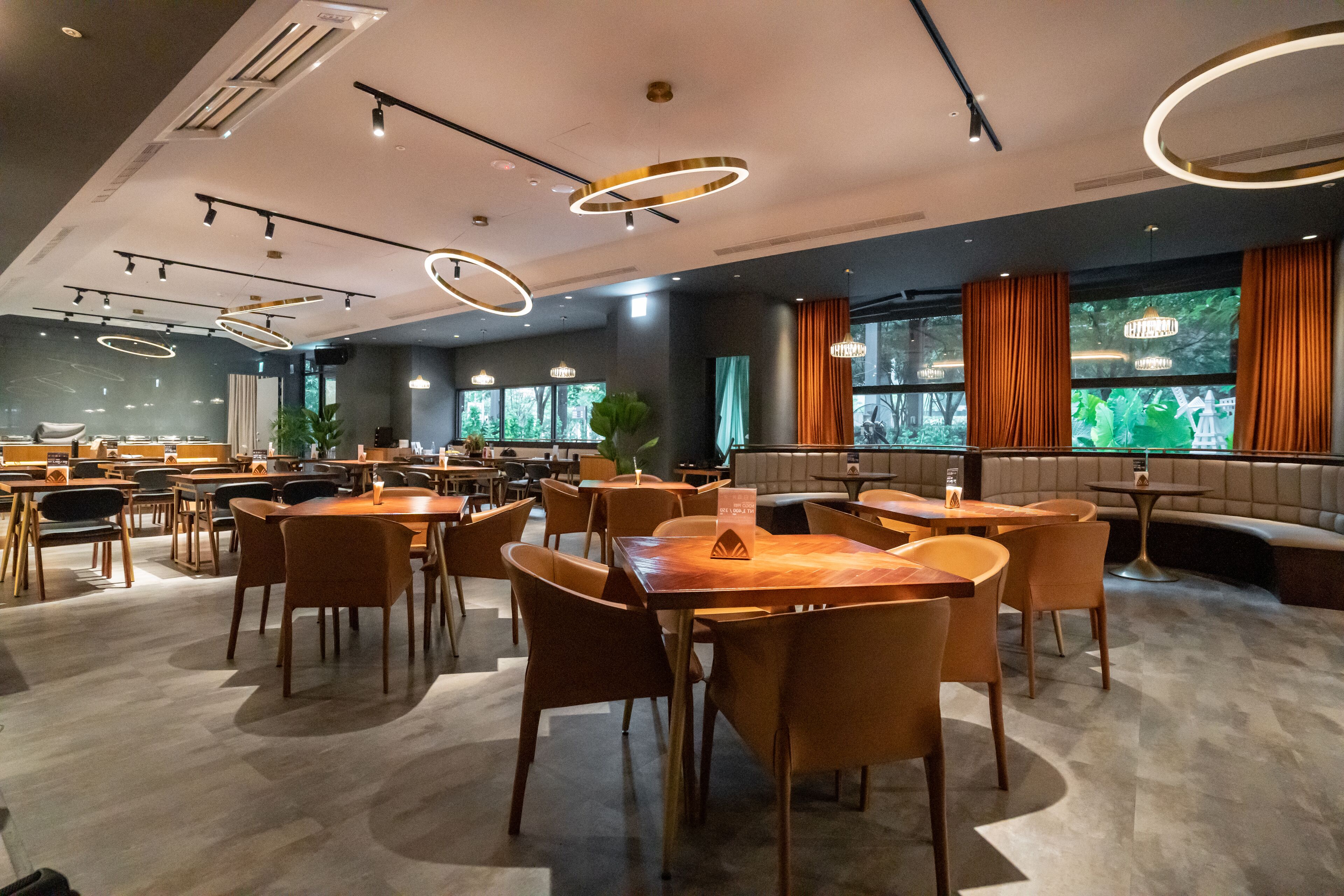 A modern dining area with multiple tables and chairs, a seating area with a sofa, and large windows with curtains.