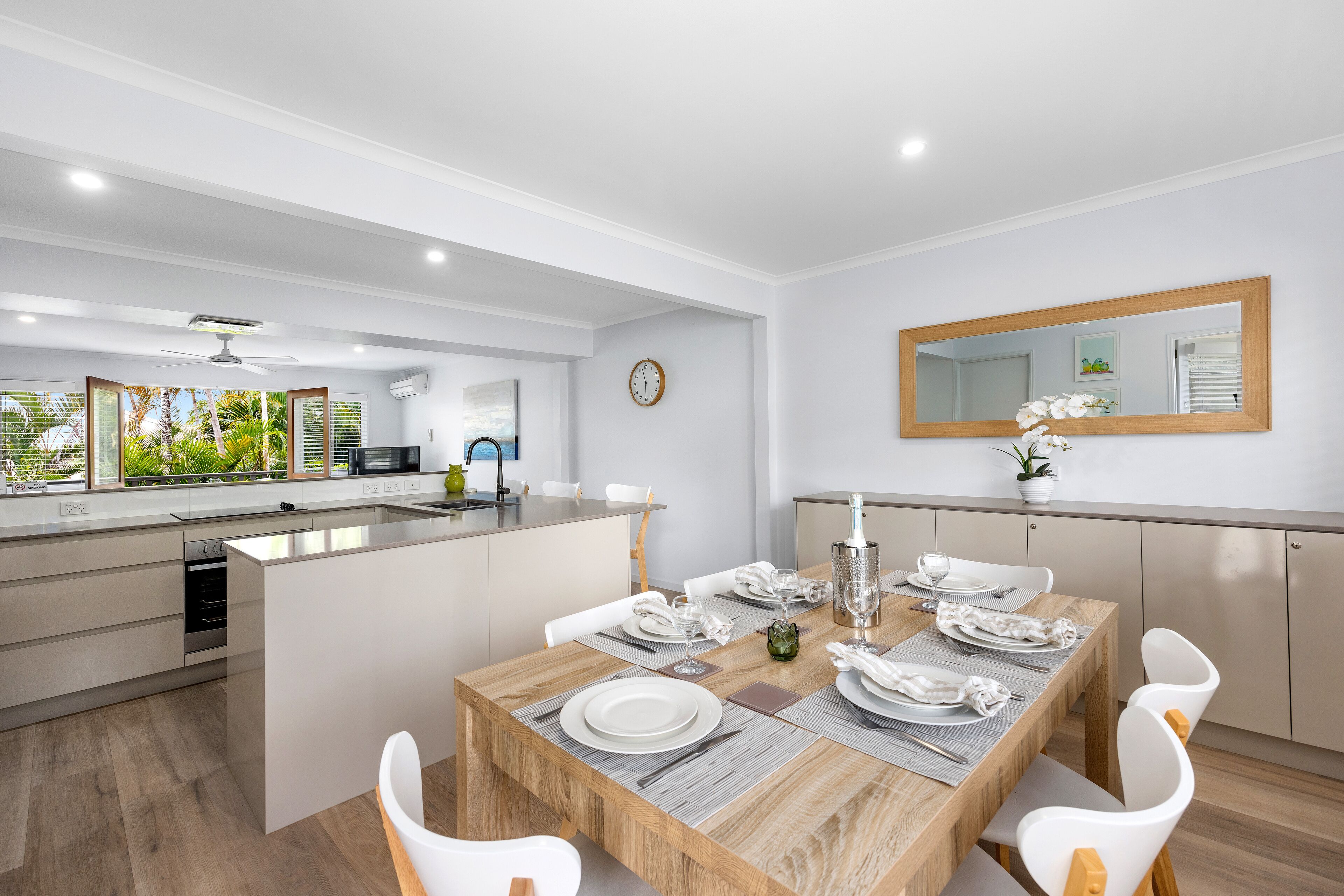 Una cucina e zona pranzo moderne con un tavolo da pranzo in legno apparecchiato per sei persone, sedie bianche e un grande specchio sopra il tavolo.
