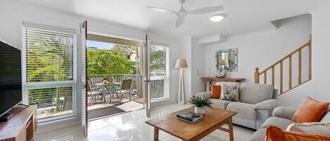 A modern living room with a sofa, coffee table, and TV stand. There is a balcony with a view of greenery.