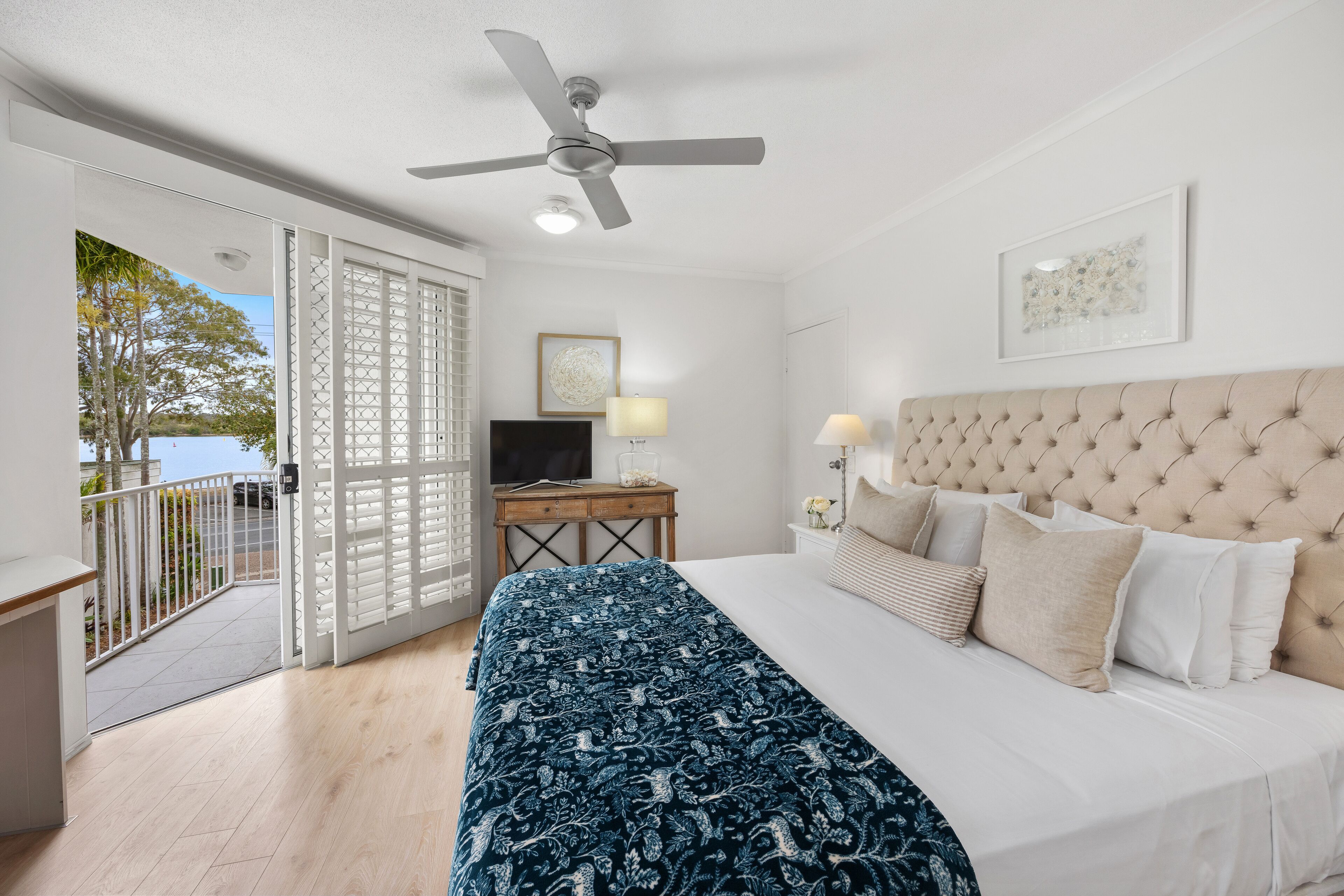 Une chambre à coucher avec un lit, un ventilateur de plafond, un bureau avec une lampe et une vue sur l’extérieur.