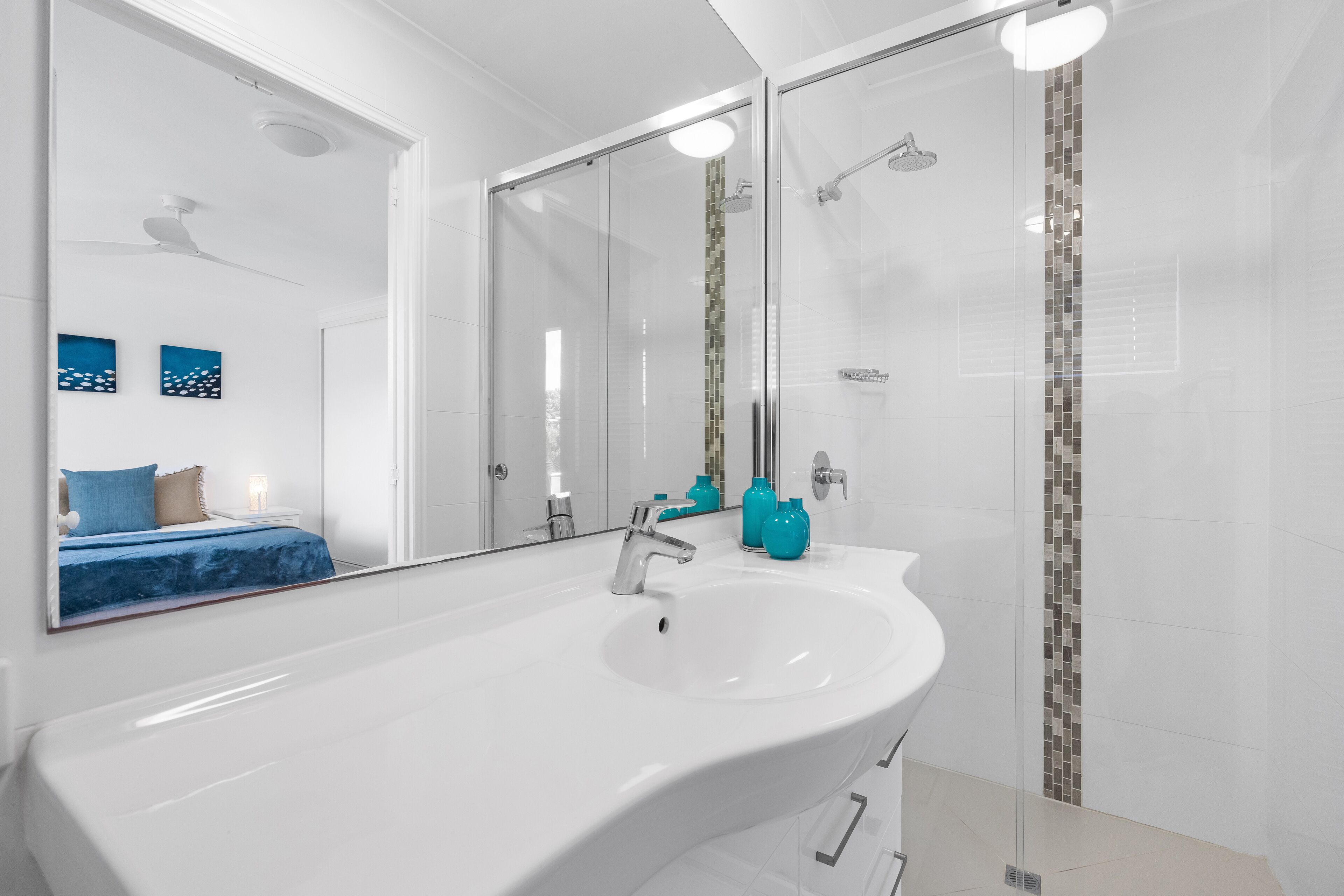 A modern bathroom with a large white sink, a glass shower enclosure, and a mirror reflecting a bed with blue bedding.