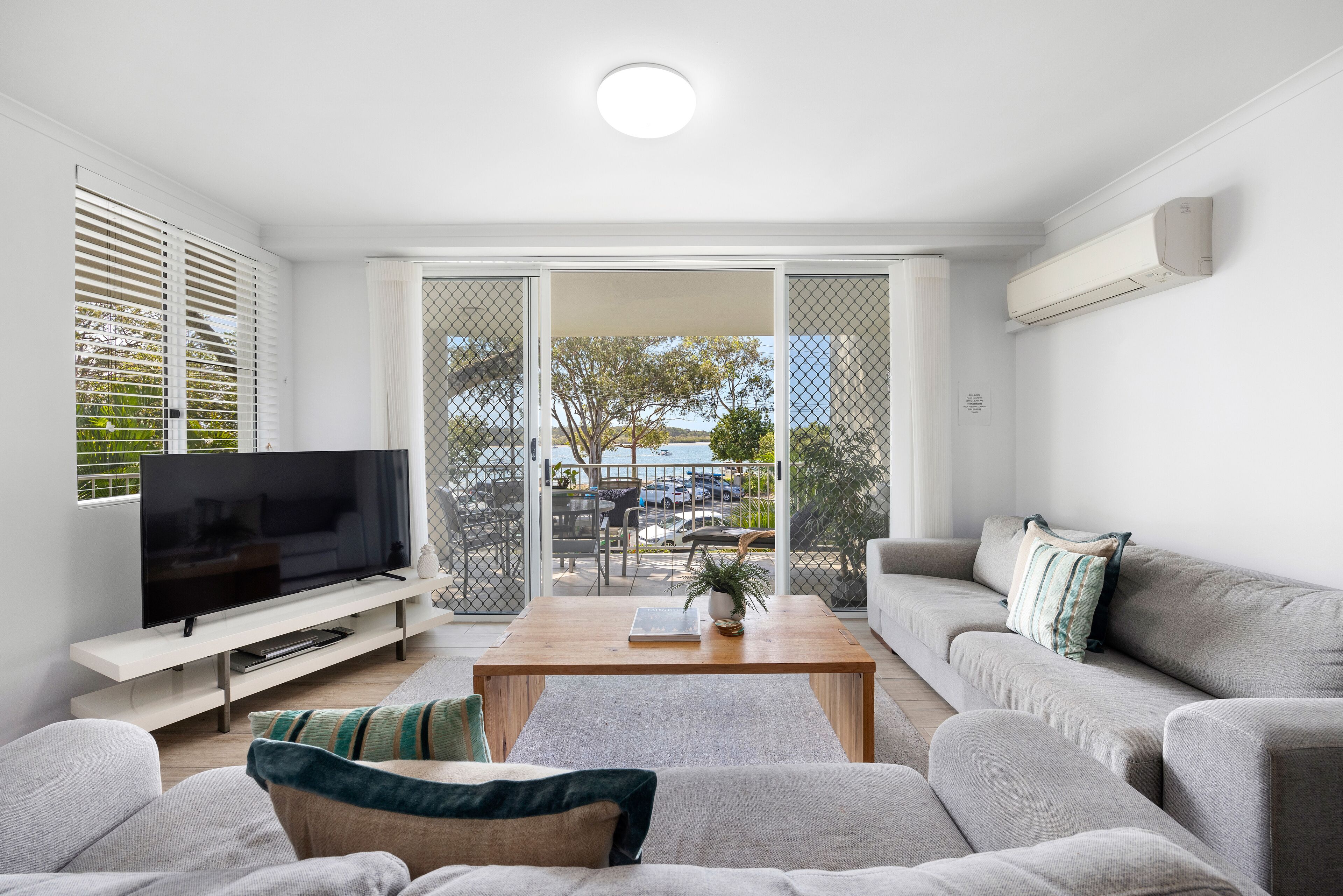 A modern living room with a flat-screen TV, grey sofas, a wooden coffee table, and a sliding glass door leading to an outdoor area.