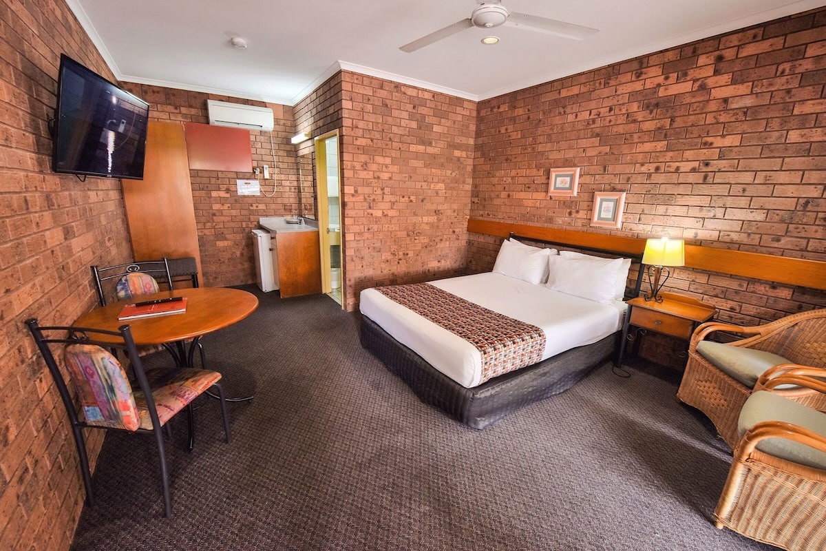 A hotel room with a brick wall, a bed, a desk, a chair, a TV, and a ceiling fan.