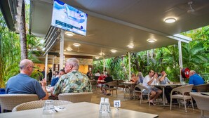 An outdoor dining area with wicker furniture, a TV screen displaying a show, and lush greenery in the background. - Knotts Crossing Resort (Katherine)