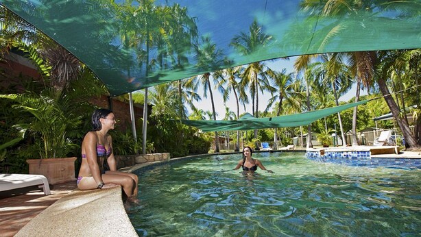 A swimming pool with clear water, surrounded by palm trees and a shaded area. - Knotts Crossing Resort (Katherine)