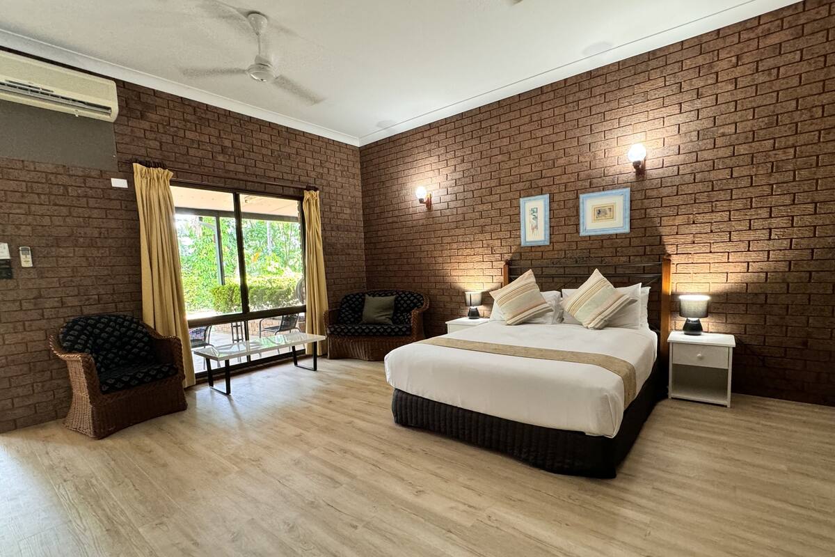 A spacious bedroom with a large bed, a sitting area with a table and chairs, and a view of greenery through a large window.