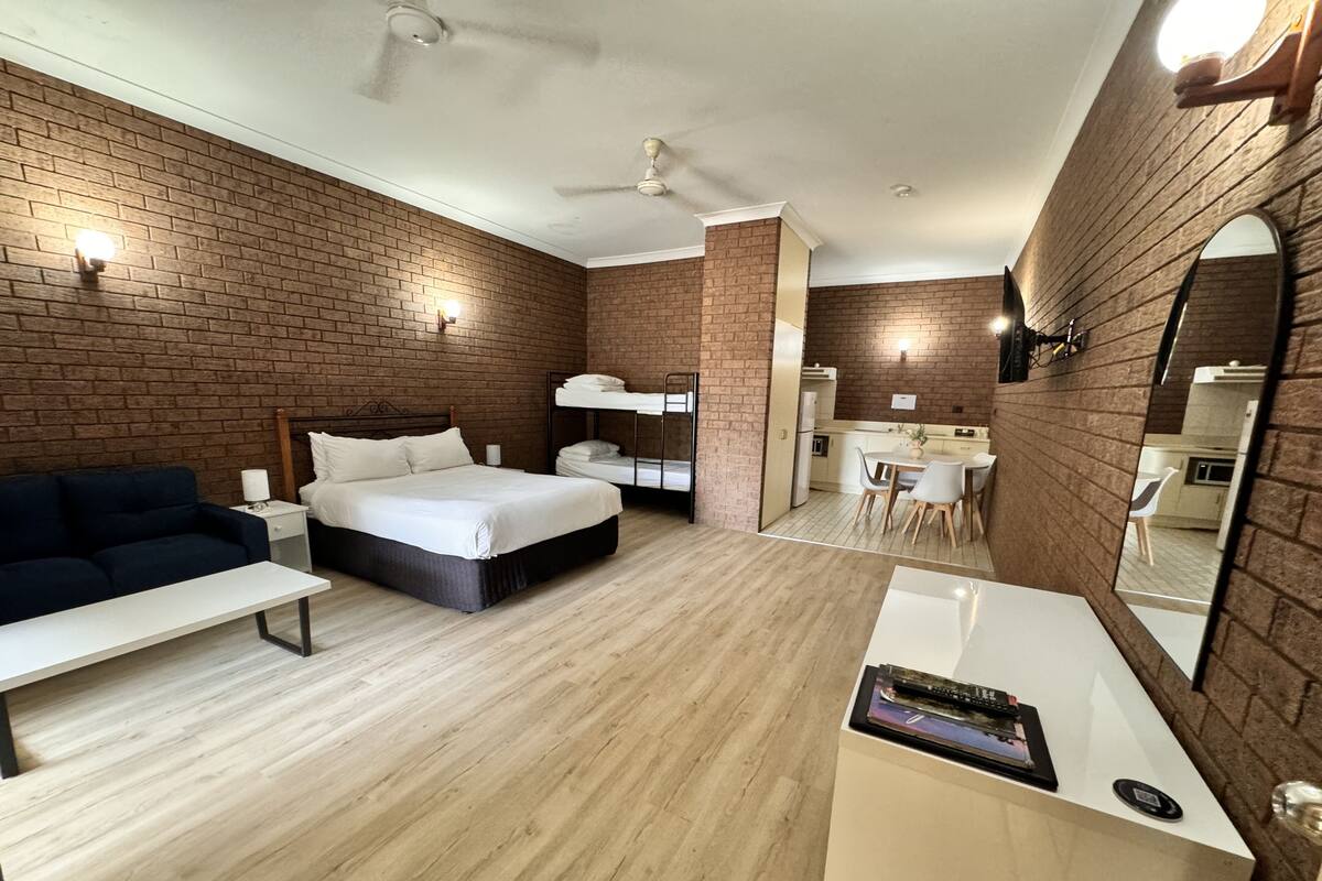 A modern hotel room with a brick wall, a bed, a desk, and a small kitchenette.