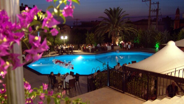 A swimming pool with a tiled deck, surrounded by outdoor seating and palm trees. - Hotel Merope (Samos)