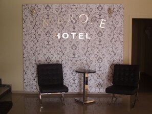 A hotel lobby with a floral wallpaper, two black upholstered chairs, and a small round table. - Hotel Merope (Samos)