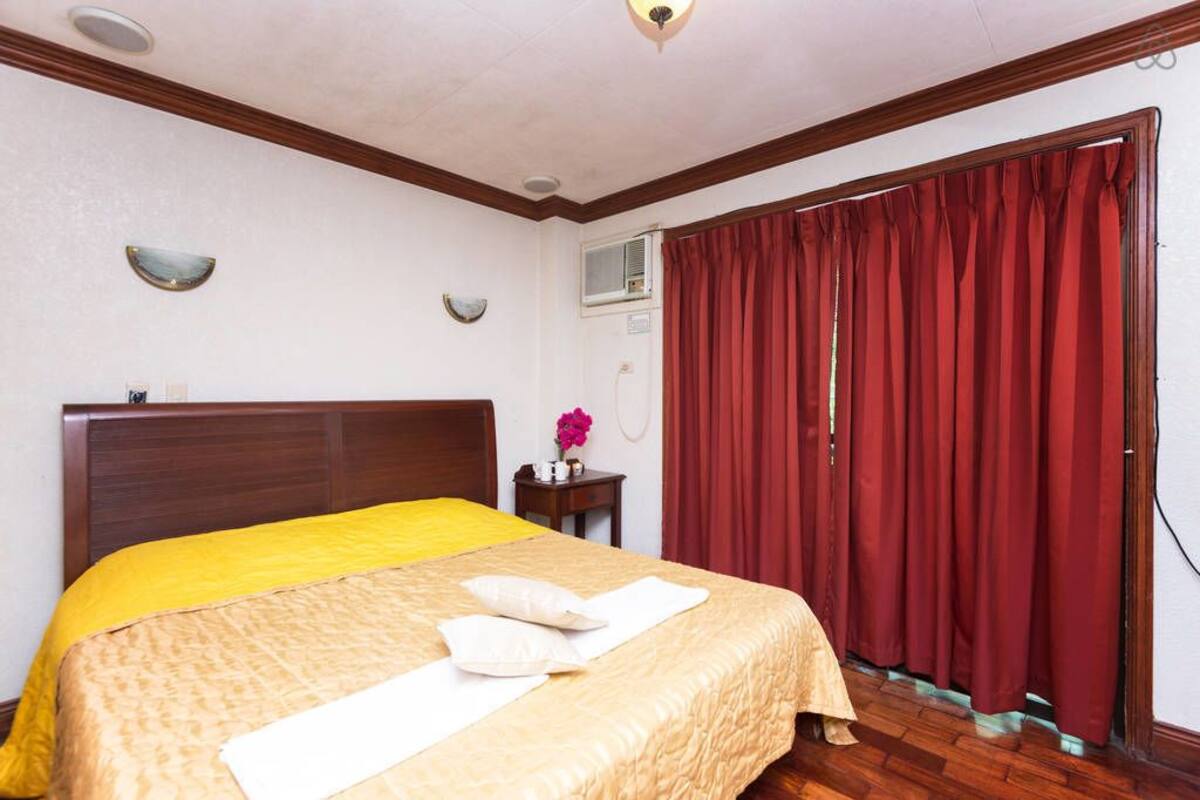 A hotel room with a bed, a nightstand, a window with red curtains, and a wall-mounted TV.
