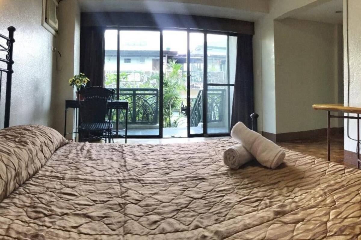 A bedroom with a large bed, a chair, and a table. There is a balcony with a view of the outside.