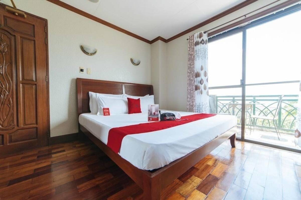 A hotel room with a wooden floor, a large bed with white bedding and a red throw, a wooden wardrobe, and a balcony with a view.