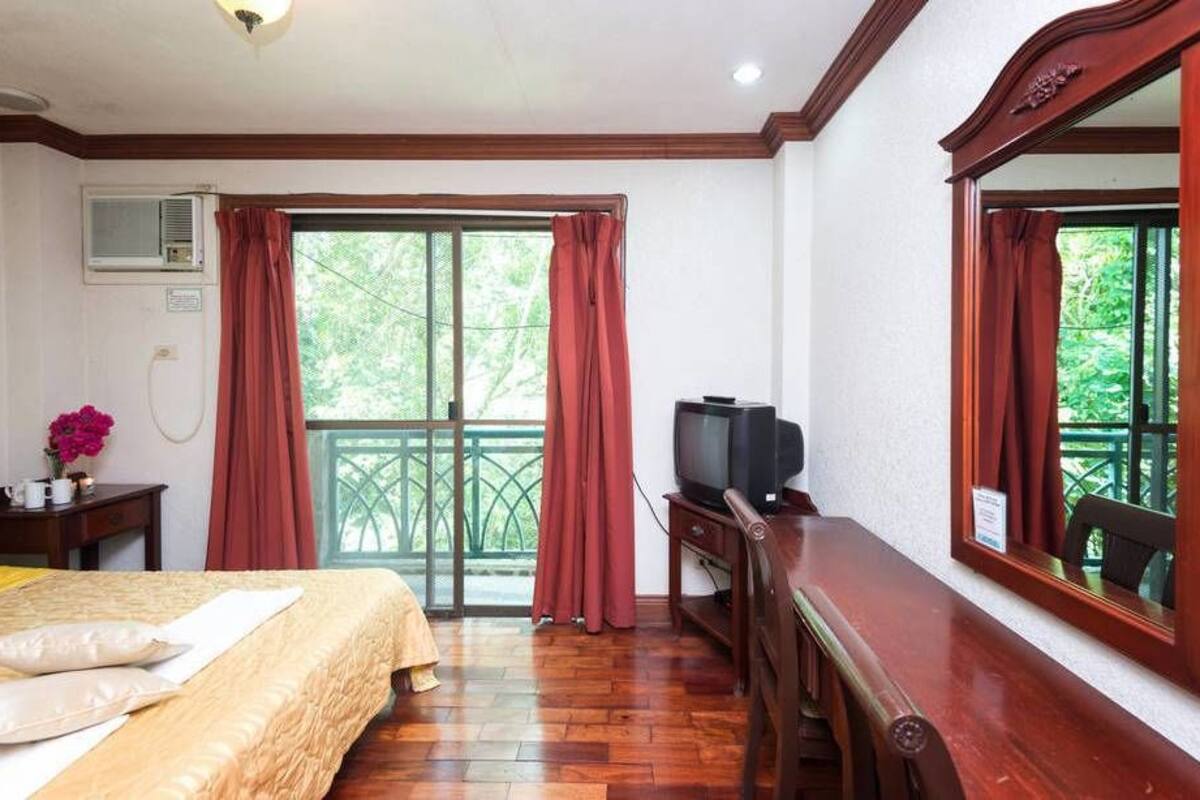 A hotel room with a bed, a television, a wooden desk, and a balcony with red curtains.