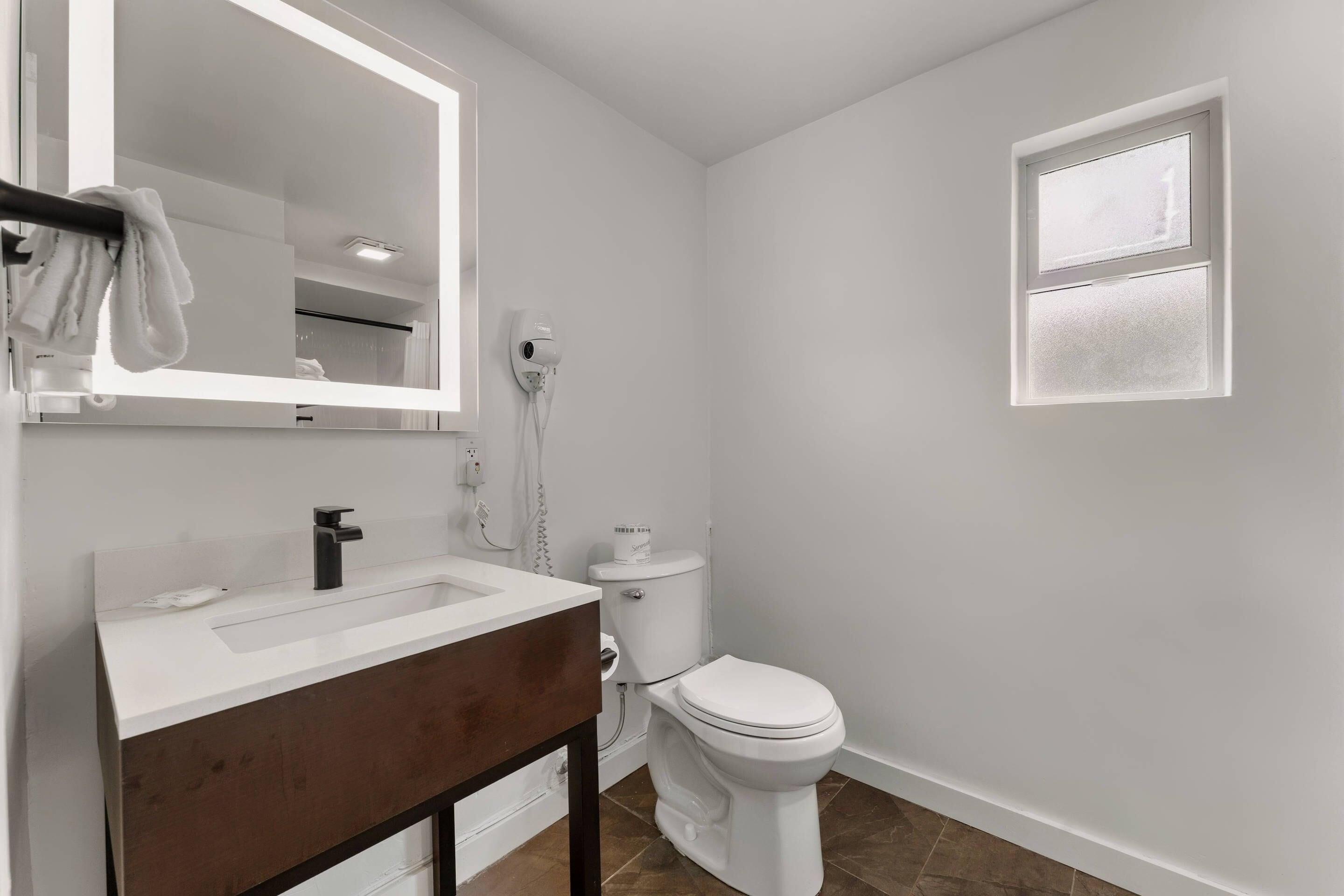 Un baño con lavamanos blanco, un espejo, inodoro y un ventanal pequeño.