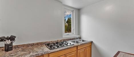 A kitchen with a gas stove, sink, wooden cabinets, a window with a view, and a countertop with utensils.