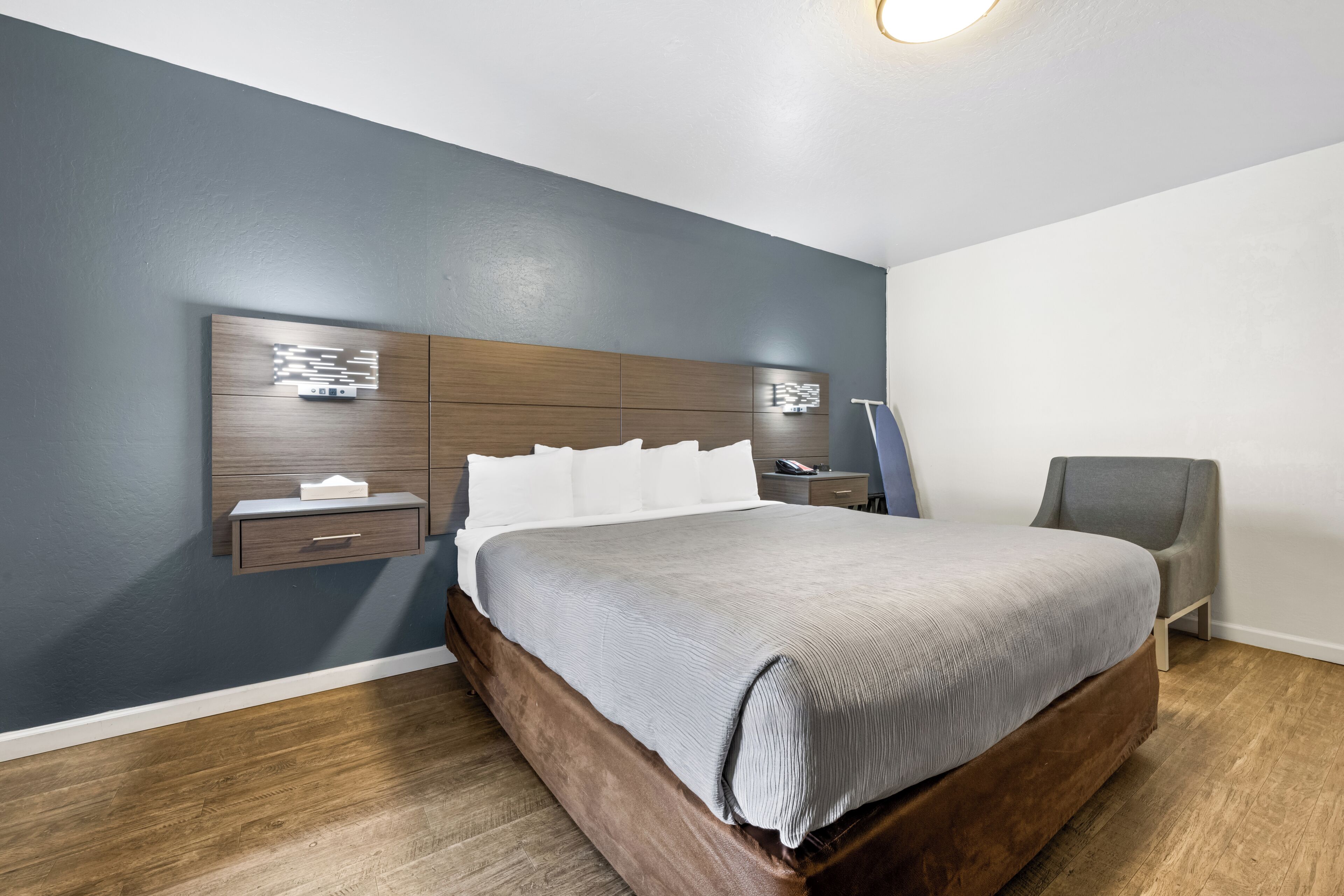 A modern bedroom with a wooden headboard, a grey bedspread, a bedside table with a lamp, and a grey armchair.